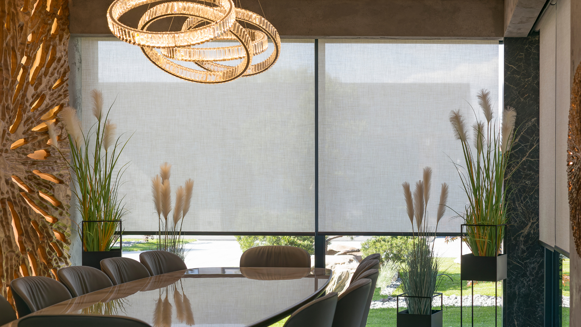 Dining room with a long table, tall plants, and window shades. A modern chandelier hangs above the table.