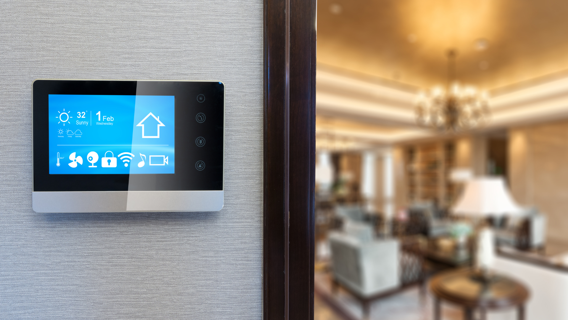 Smart home control panel on a wall, with a living room visible through a doorway. Blue display, silver trim.