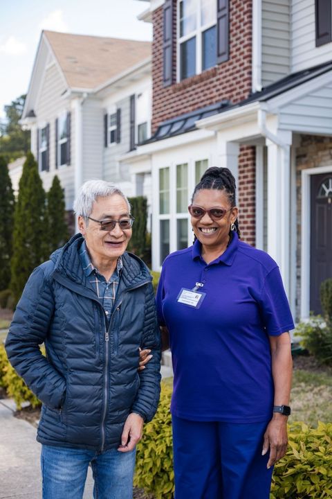 Caregiver in blue scrubs assists older person outdoors, smiling.