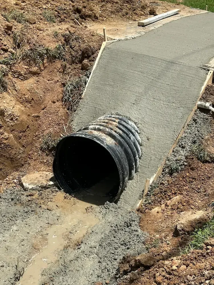 A concrete drainage culvert installed under a new concrete driveway, brown dirt and green grass surround it.