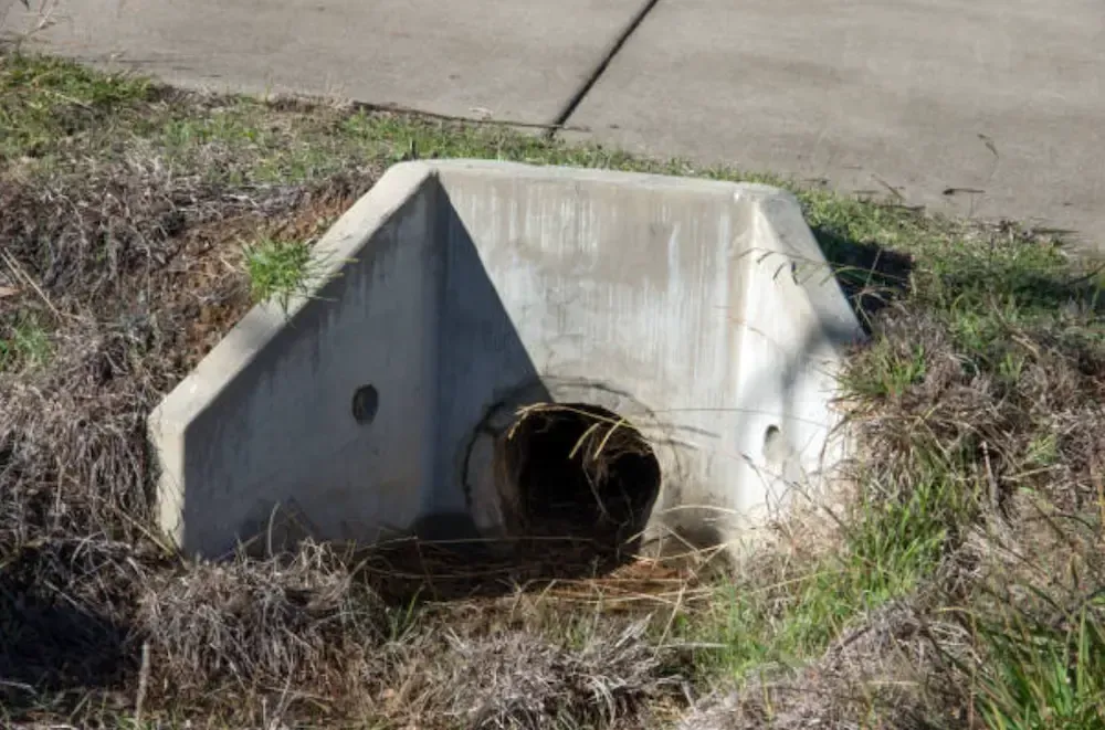 Concrete culvert with a circular opening in a grassy area next to a sidewalk.
