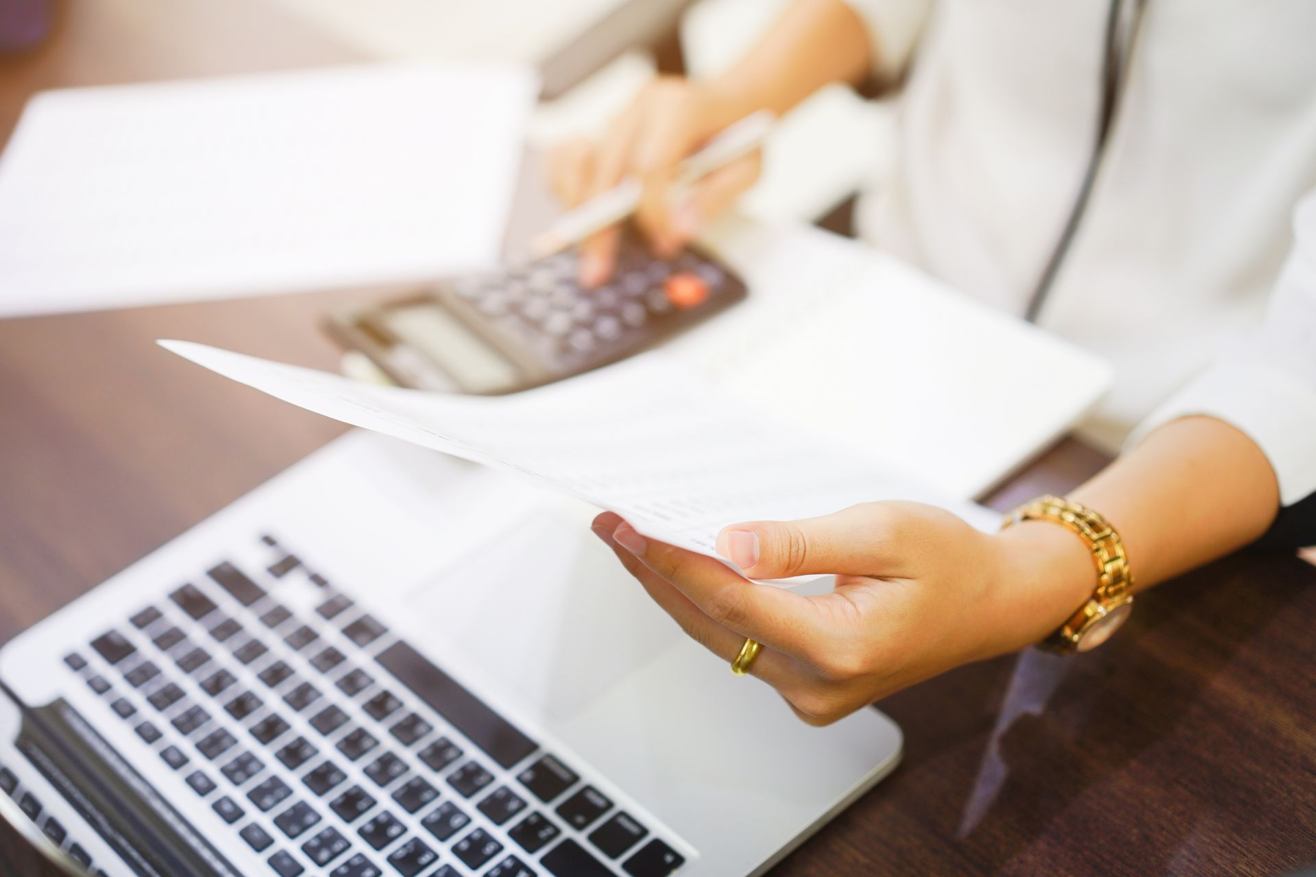 A person using a calculator, looking at papers, and working on a laptop at a desk.