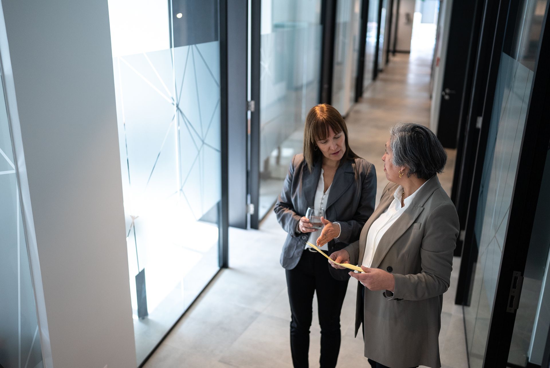 Two women in suits talking in a bright hallway with glass walls.