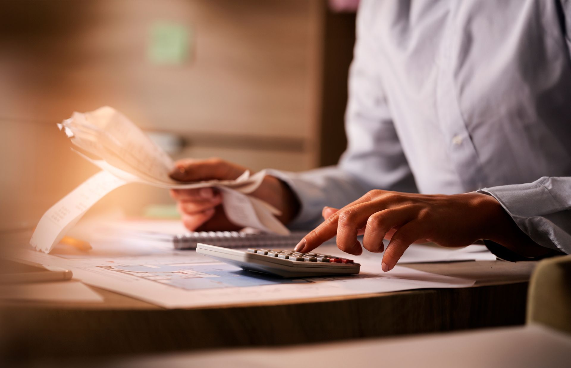 Person calculating bills with a calculator, documents on desk, indoors, soft lighting.