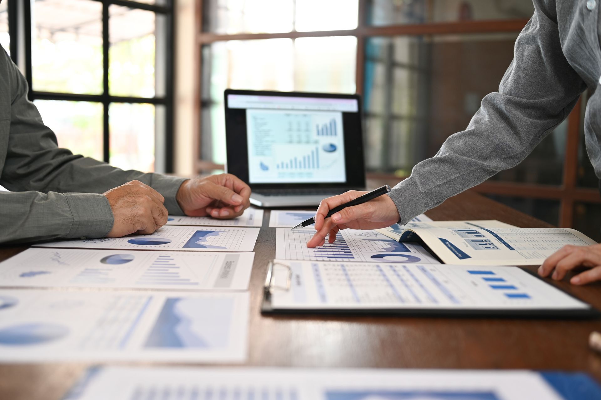 Two people reviewing financial reports at a table, laptop open with charts displayed, pointing and discussing data.