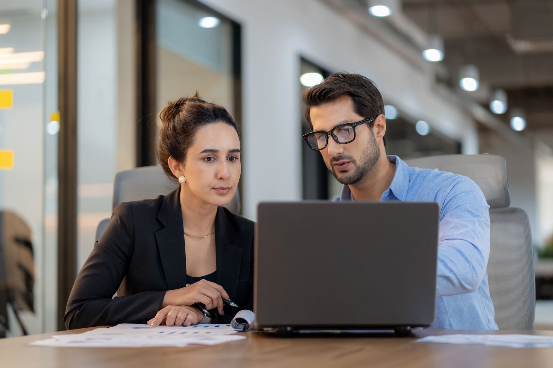 A woman and a man are looking intently at a laptop screen in a modern office setting.