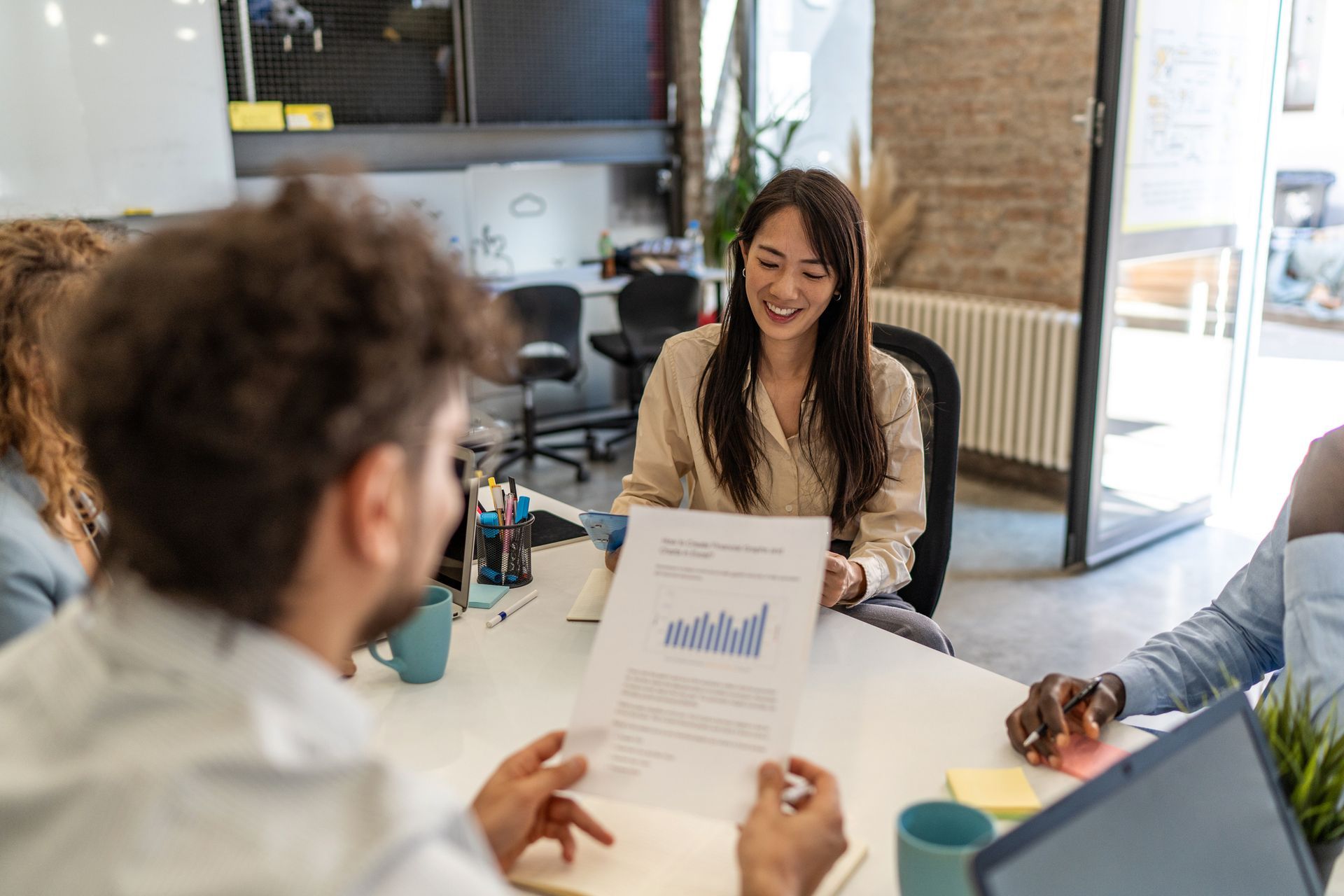 Business meeting with diverse colleagues at a round table. A woman smiles, holding a report with a blue graph.