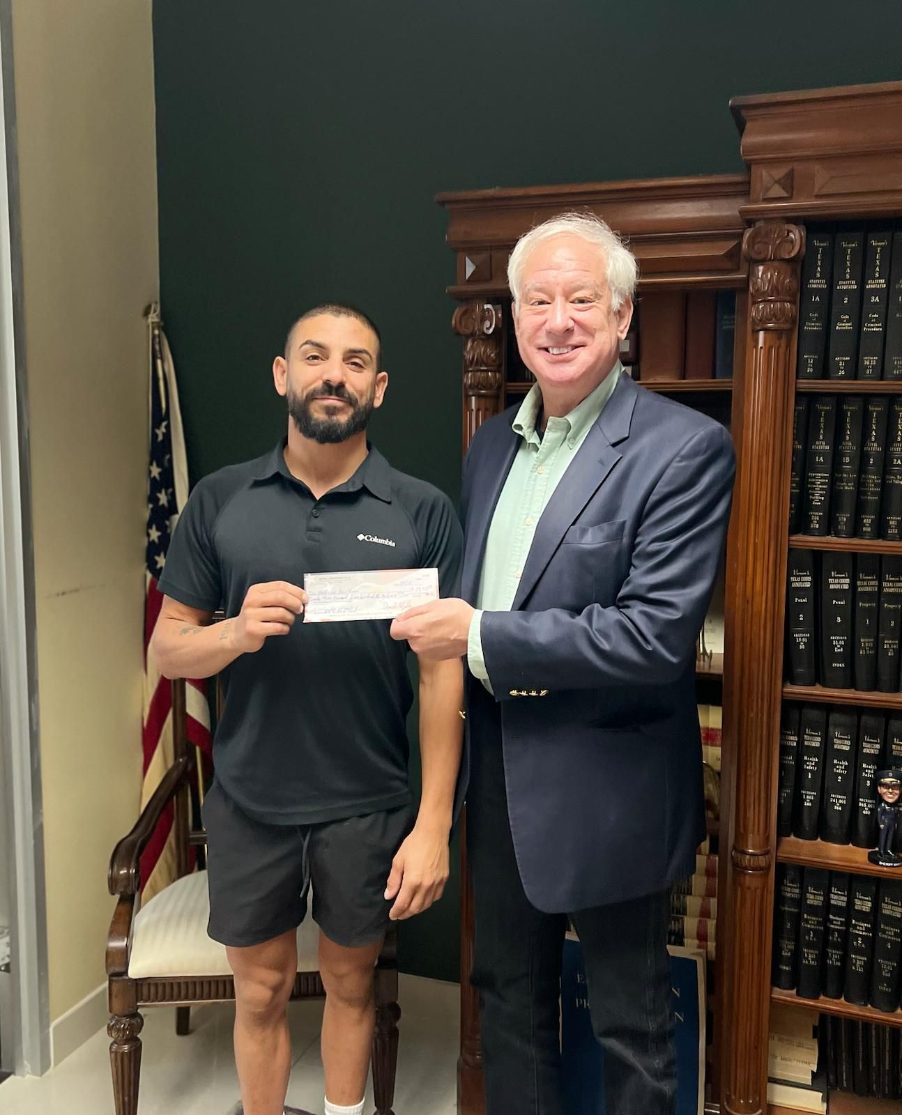 Two men, one in black, the other in a suit, in a room with bookshelves and an American flag, one handing a check.