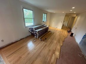 A room under renovation with partially installed light-wood flooring, a stack of wood planks, and unfinished walls.