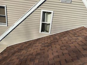 High-angle view of a brown asphalt shingle roof adjacent to a house with beige horizontal siding and two windows.