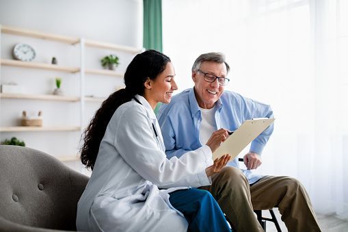 A doctor is sitting on a couch with an elderly man and looking at a clipboard.