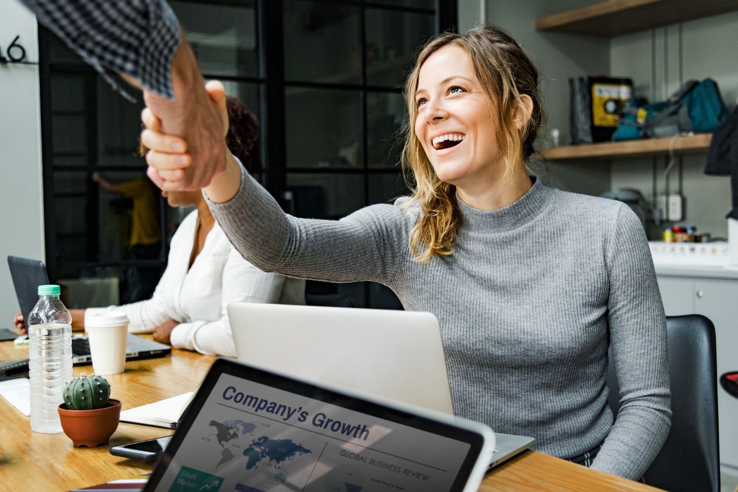 Woman in grey shirt shakes hands, smiling at a business meeting. Other person, laptops, and office setting.