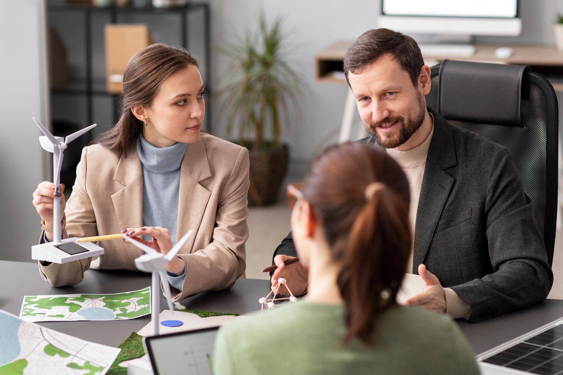 Three people in an office discuss renewable energy models, including wind turbines and solar panels.
