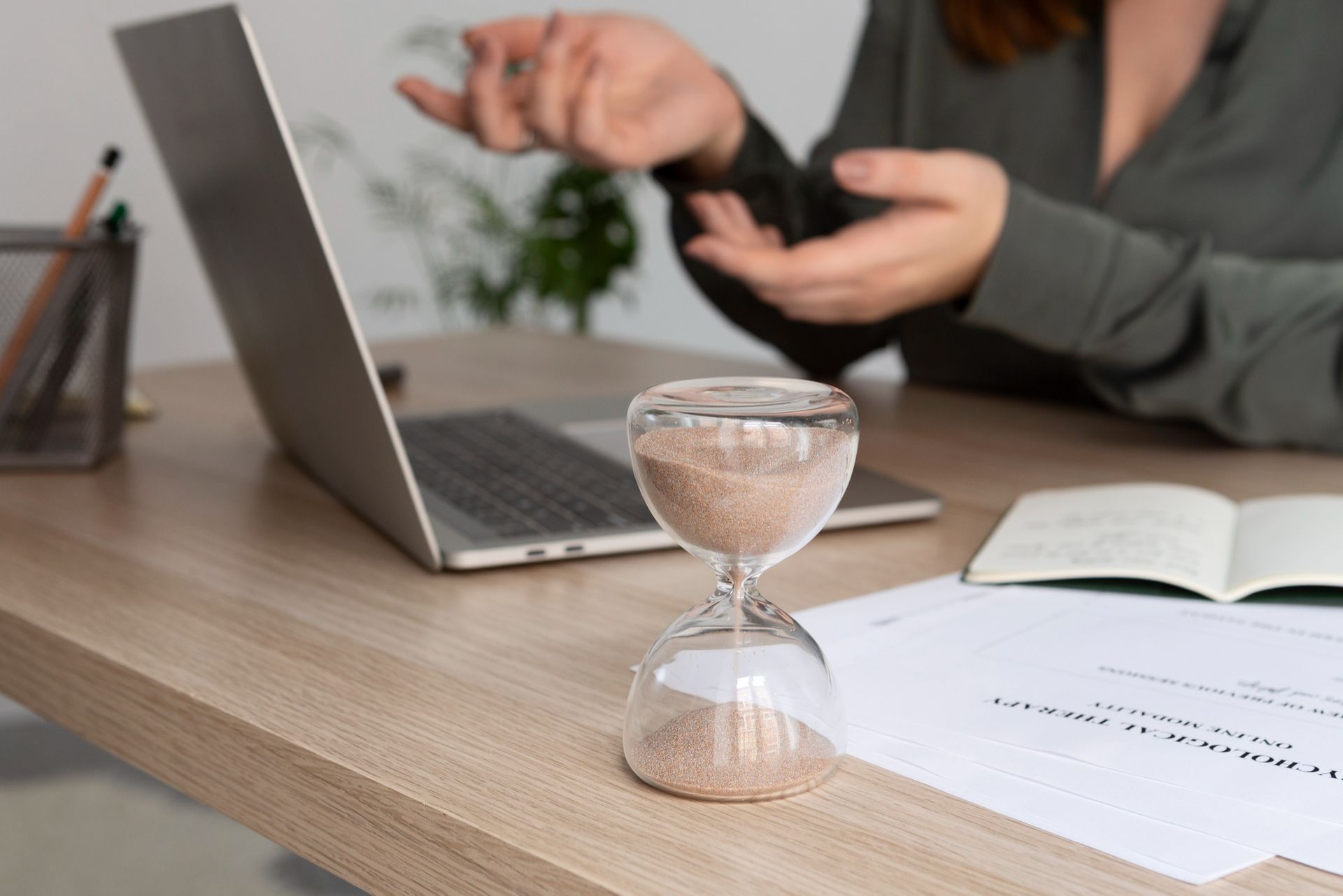 Hourglass on desk with laptop and papers; person gesturing in background.