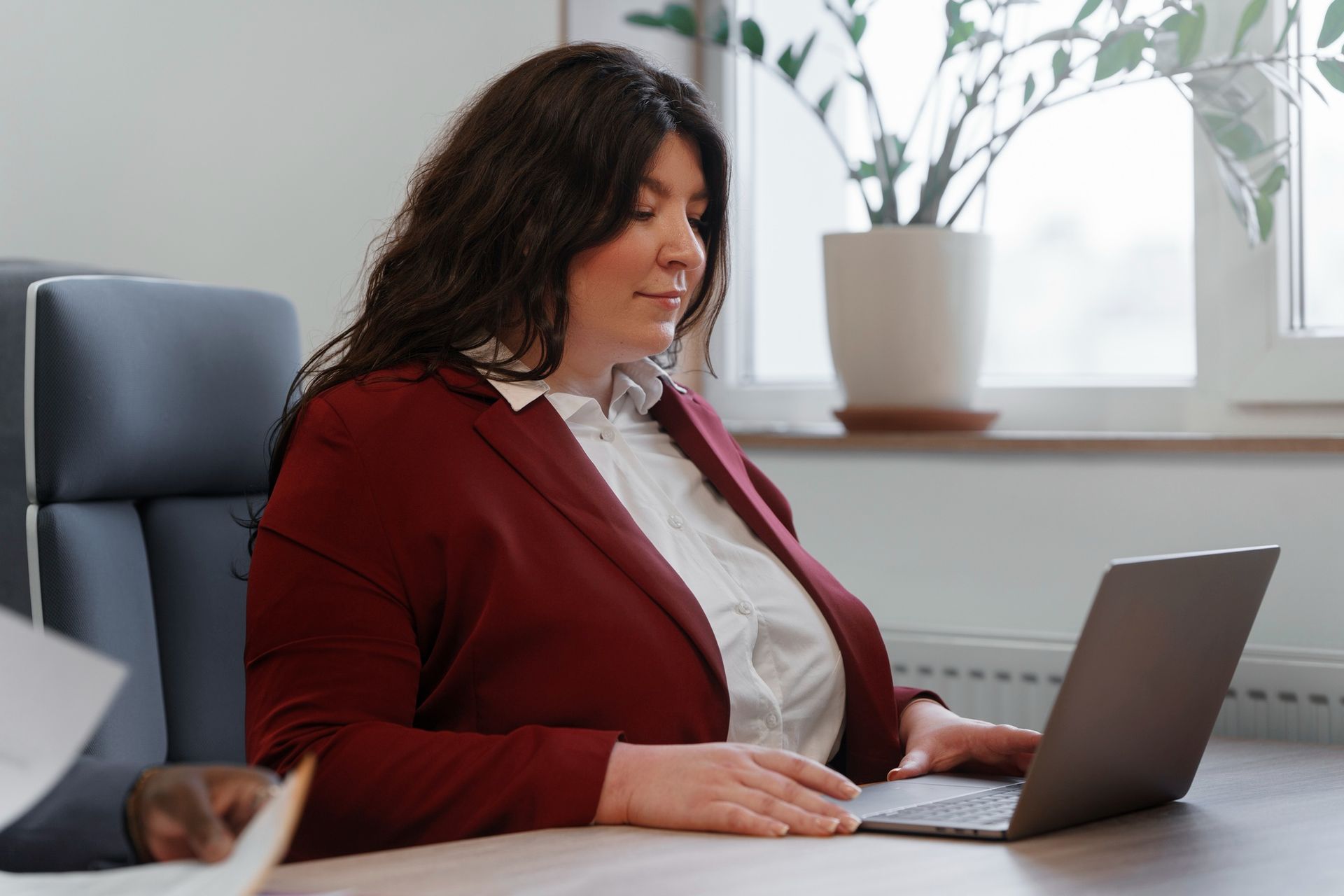 Woman in a red blazer working on a laptop at a desk in a bright office