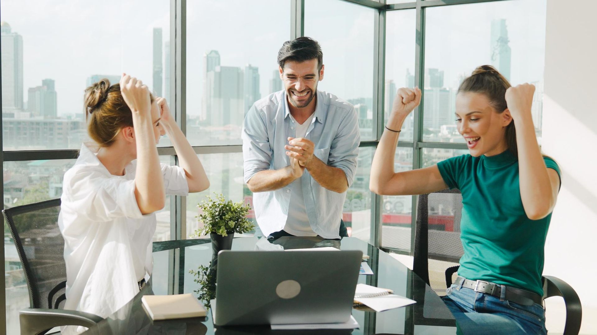 Three people celebrate around a laptop in a brightly lit office; two raise their arms in excitement, the other claps.