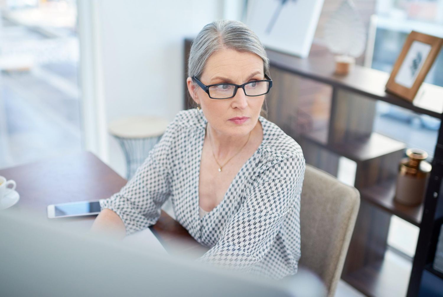 Woman in glasses working at a desk in a bright office, looking to the side with a thoughtful expression