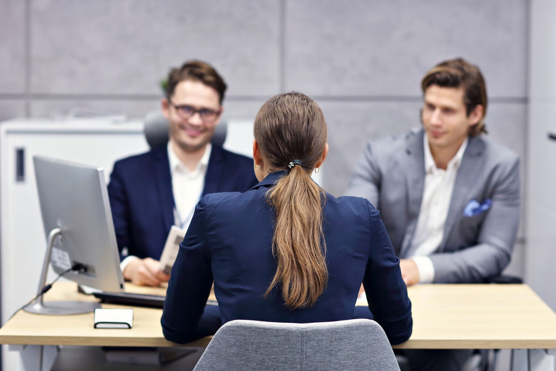 Woman in interview facing two men in suits, sitting at desk in office.