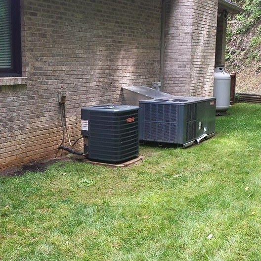 Two air conditioning units next to a brick building on a grassy lawn. A propane tank is in the background.