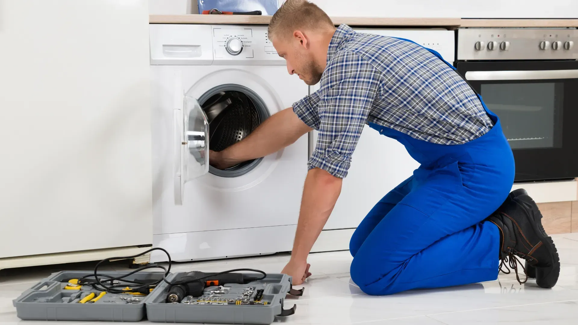 A man is kneeling on the floor fixing a washing machine.
