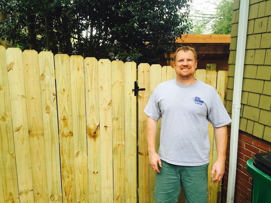 Man standing in a backyard beside a wooden fence and green trash bin