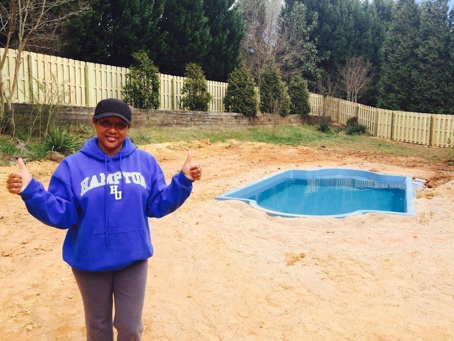 Person in blue sweatshirt giving thumbs up beside a backyard pool and sandy patio.
