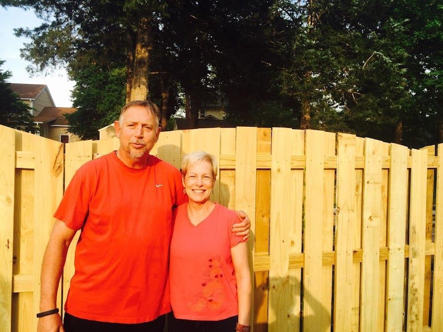 Two people standing together in front of a wooden fence in a sunny yard, both wearing red shirts.