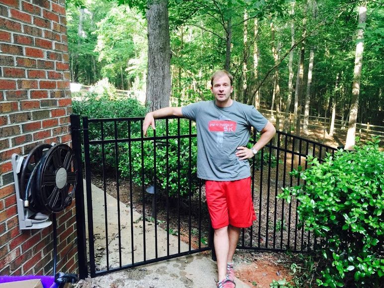 Man leaning on a black gate beside a brick house, wearing a gray T-shirt and red shorts in a wooded yard