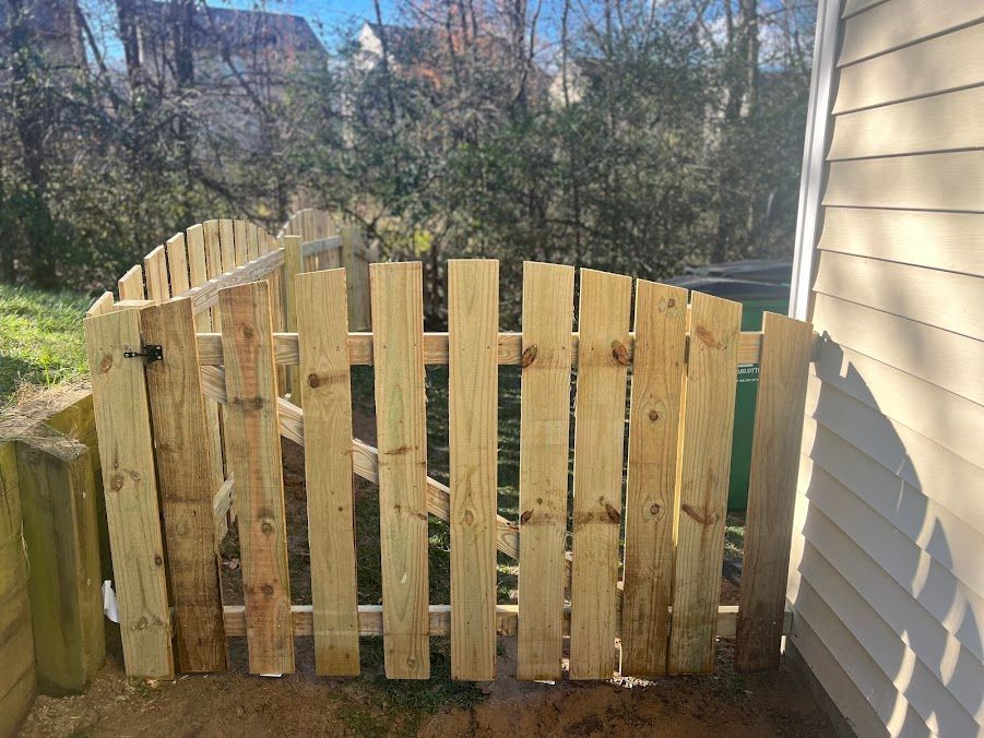 Weathered wooden gate in a rustic fence beside a beige house, with trees in the background.