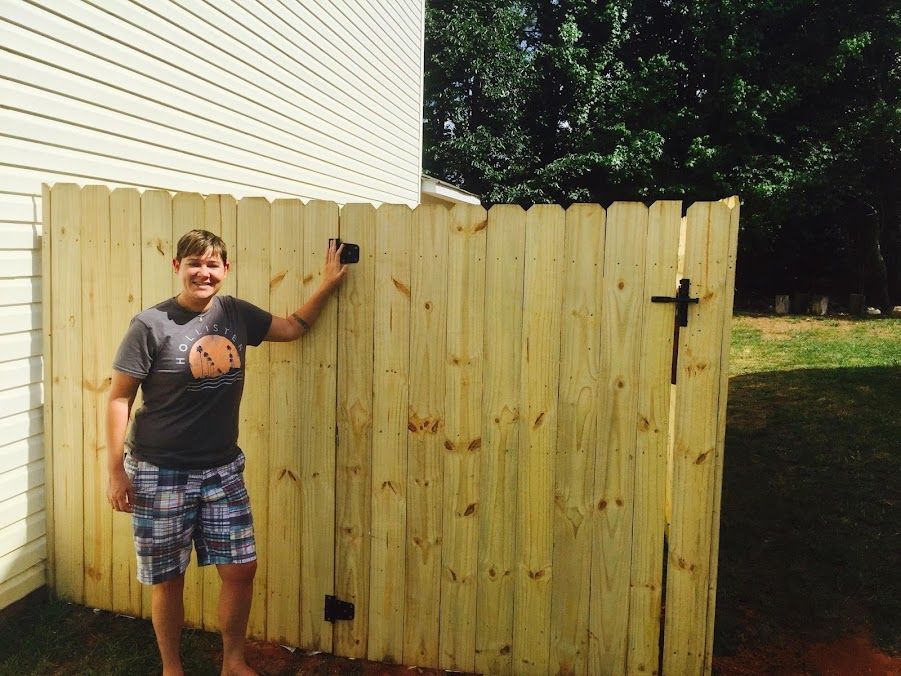 Person standing by a wooden fence gate beside a house, holding the latch and smiling outdoors.