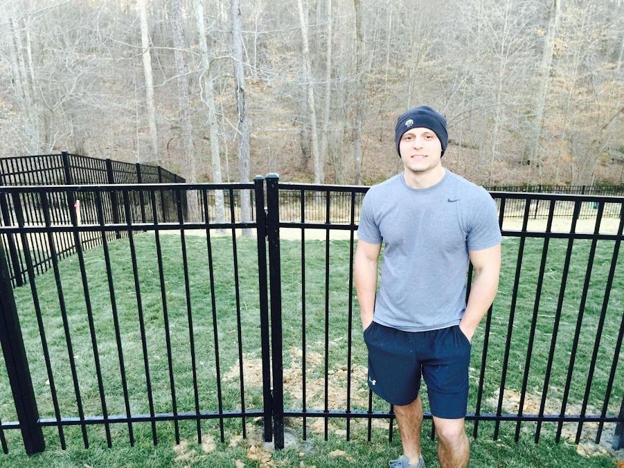 Man standing by a black metal fence in a wooded park, wearing a gray shirt and dark shorts