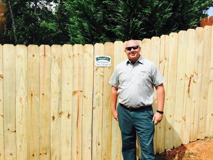 Man standing in front of a wooden fence on a sunny day, wearing sunglasses and a gray shirt.