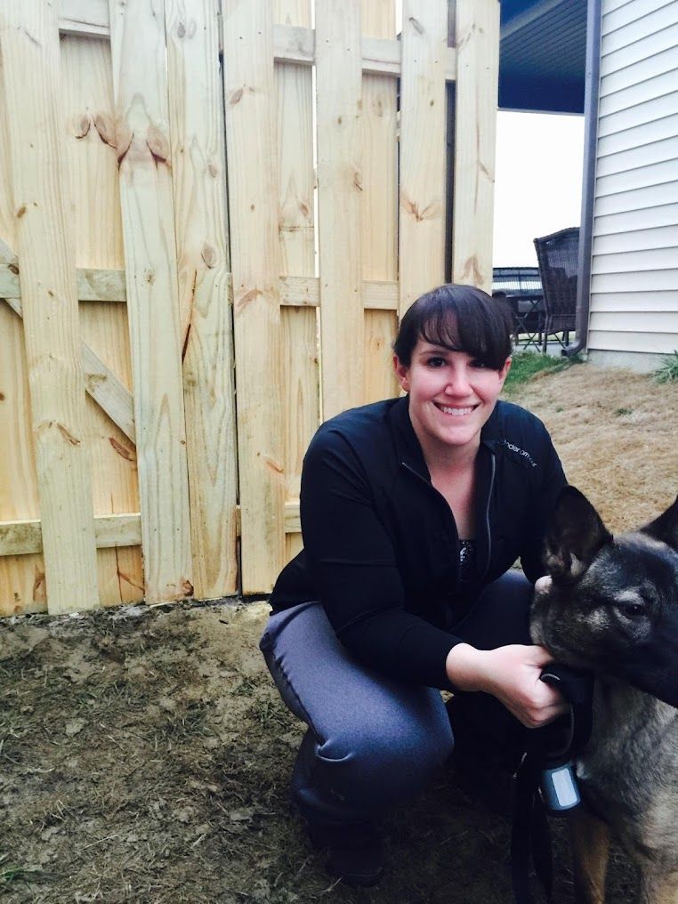 Woman kneeling outdoors beside a black dog in front of a wooden fence, smiling at the camera