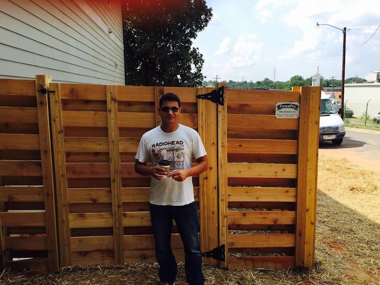 Person standing by a wooden gate, holding a drink, with a white house and street in the background