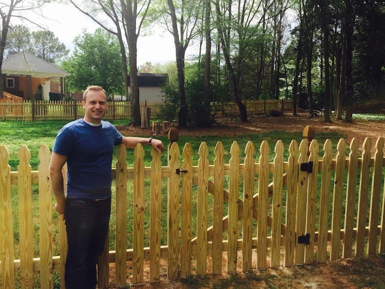 Man standing by a wooden fence in a wooded yard with houses in the background