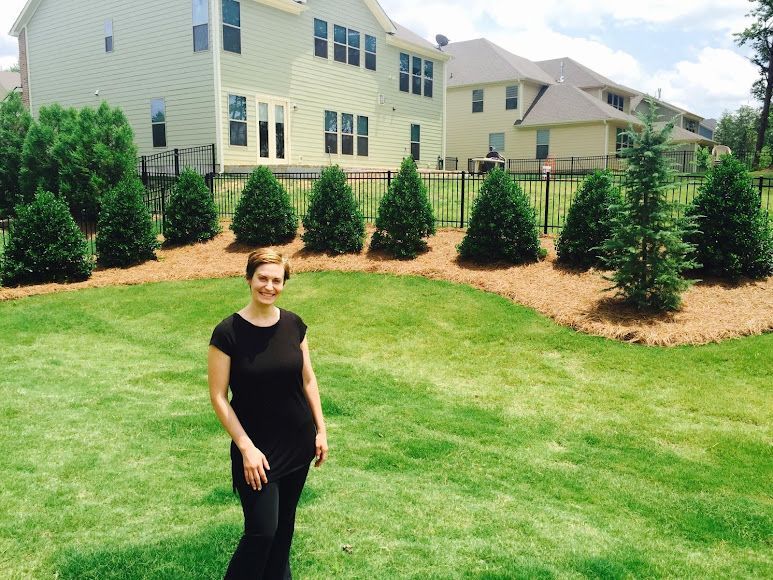 Woman standing on a grassy lawn in front of a beige house and row of shrubs.