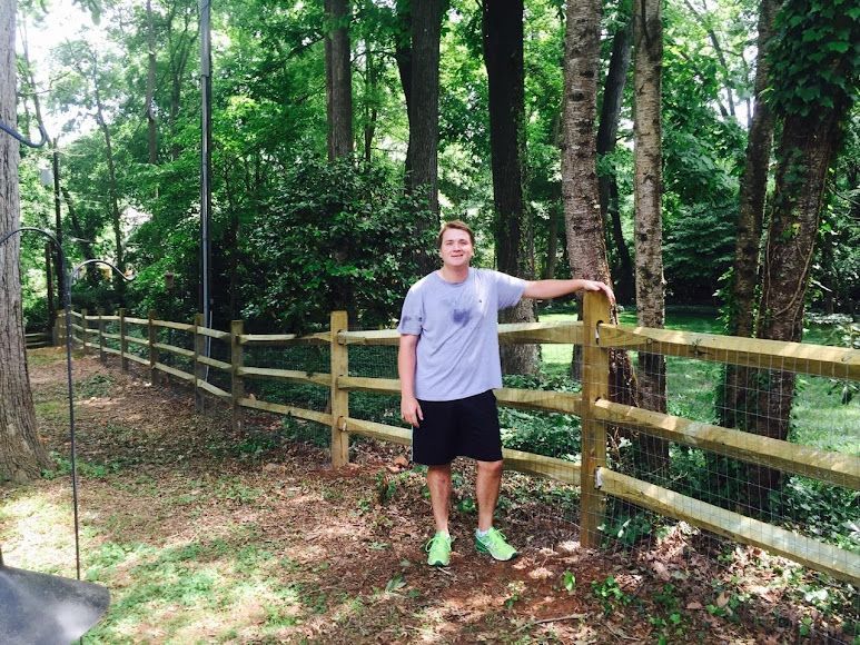 Person standing by a wooden fence on a tree-lined trail, wearing a gray shirt and black shorts.