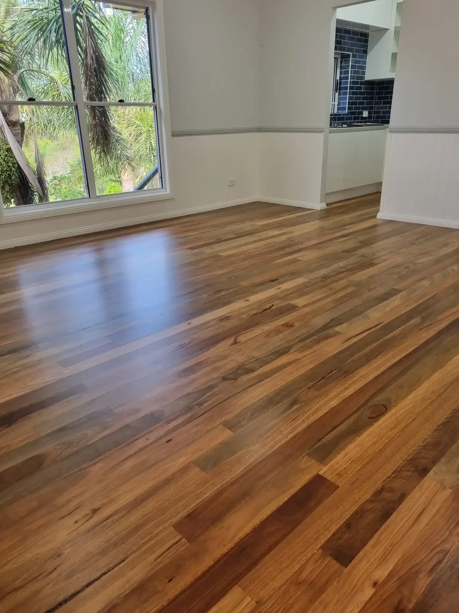 Wooden floor reflecting light near a window overlooking trees. A doorway leads to a kitchen. — The Good Wood Polished Timber Floors in Gatton, QLD