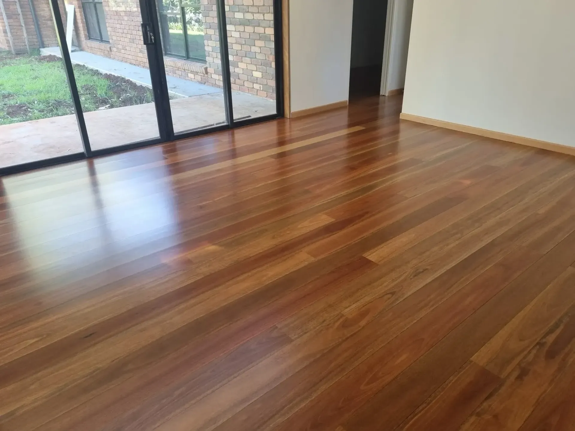 Polished hardwood floor reflecting light; a room with glass sliding doors and a white wall. — The Good Wood Polished Timber Floors in Oakey, QLD