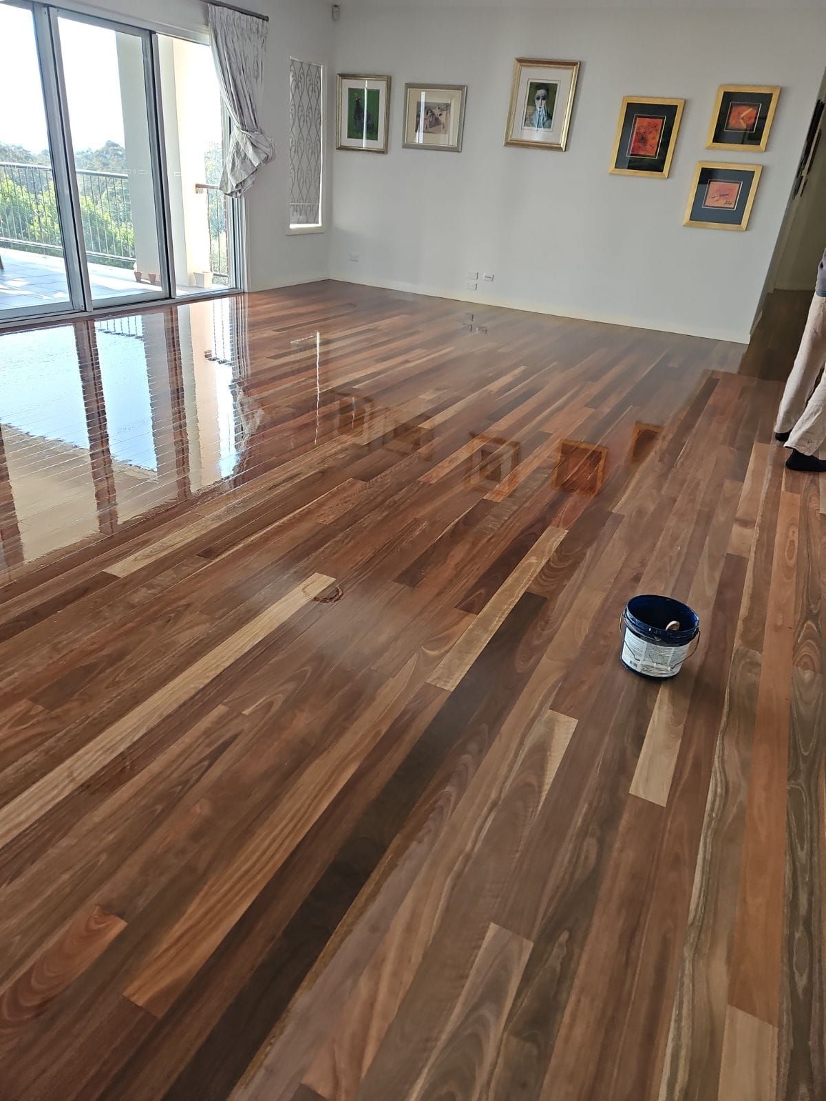 Newly finished hardwood floor reflecting light with a paint can nearby, in a bright room with artwork. — The Good Wood Polished Timber Floors in Oakey, QLD