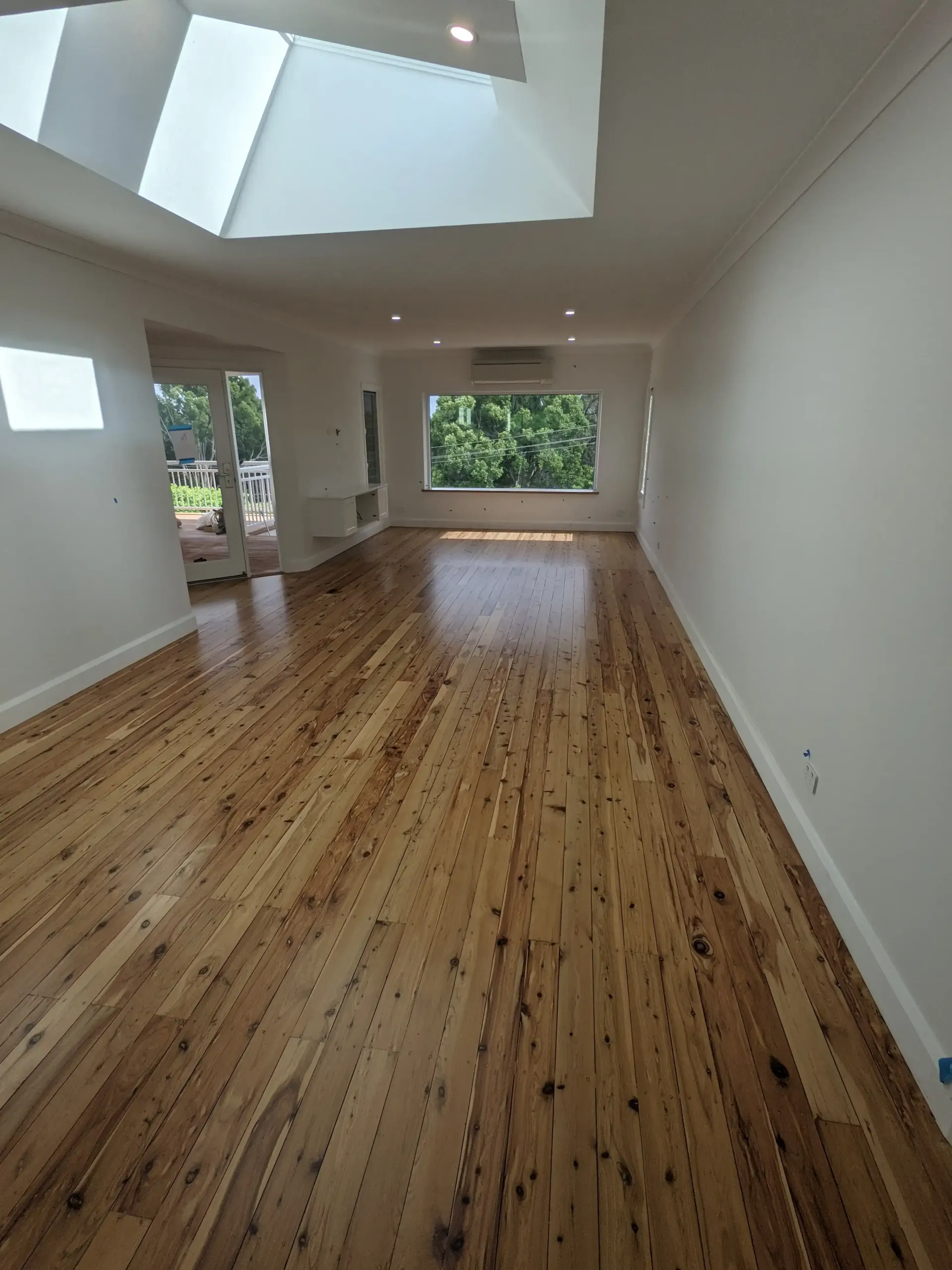 Bright living room with glossy wooden floor, white walls, large window with trees outside, and skylight. — The Good Wood Polished Timber Floors in Oakey, QLD