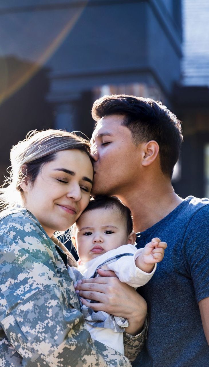 A service member in camouflage uniform holds a baby while their partner kisses their forehead outdoors in sunlight.
