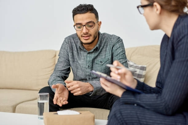 A man is sitting on a couch talking to a woman who is holding a clipboard.