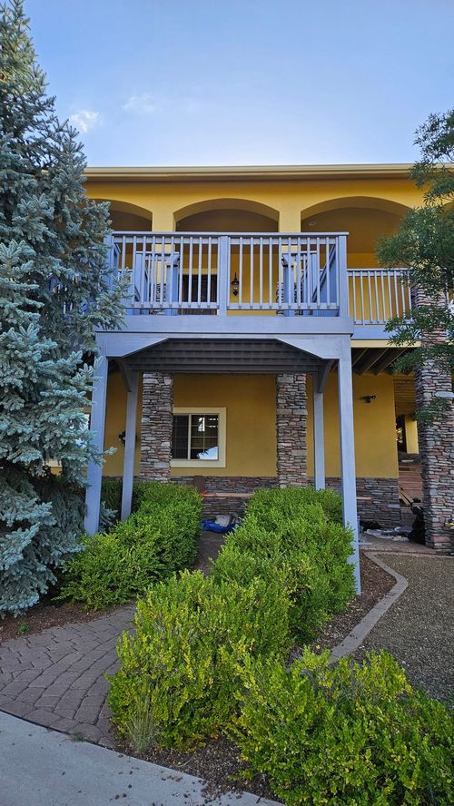 A yellow two-story building with a raised wooden balcony, stone pillars, and a brick walkway surrounded by green bushes.