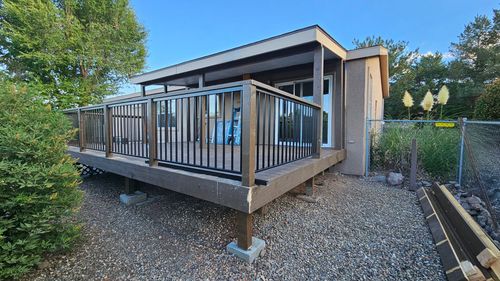 A brown elevated deck with black metal railings attached to a stucco-sided home, set on a gravel yard with nearby trees.