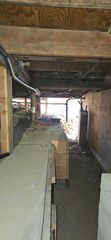 View into a dimly lit crawlspace with wooden floor joists, a dirt floor, and a narrow passage leading to an outdoor opening.