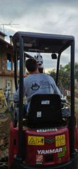A person operates a red Yanmar excavator on a construction site near a house.