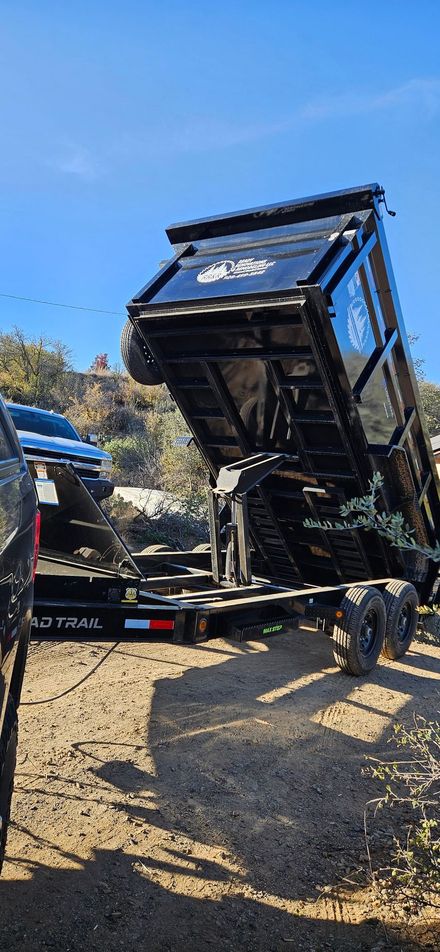 A black dump trailer hitched to a vehicle is fully raised in a gravel-filled lot under a clear blue sky.