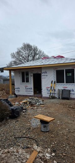 A small, unfinished house with white siding under construction, featuring a gravel yard and a wooden tool bench in the front.