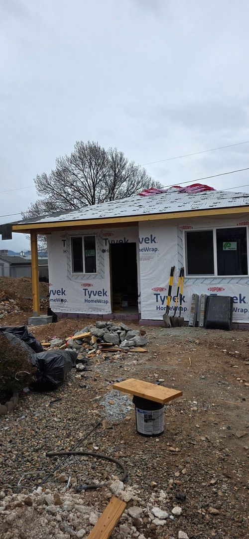 A small, unfinished house with white siding under construction, featuring a gravel yard and a wooden tool bench in the front.